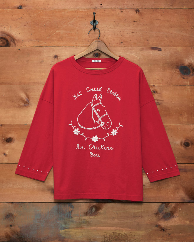 The Stables Long Sleeve Tee, crafted from 100% cotton, features a red long-sleeve raglan design with a horse graphic and white text, displayed against a wooden wall.