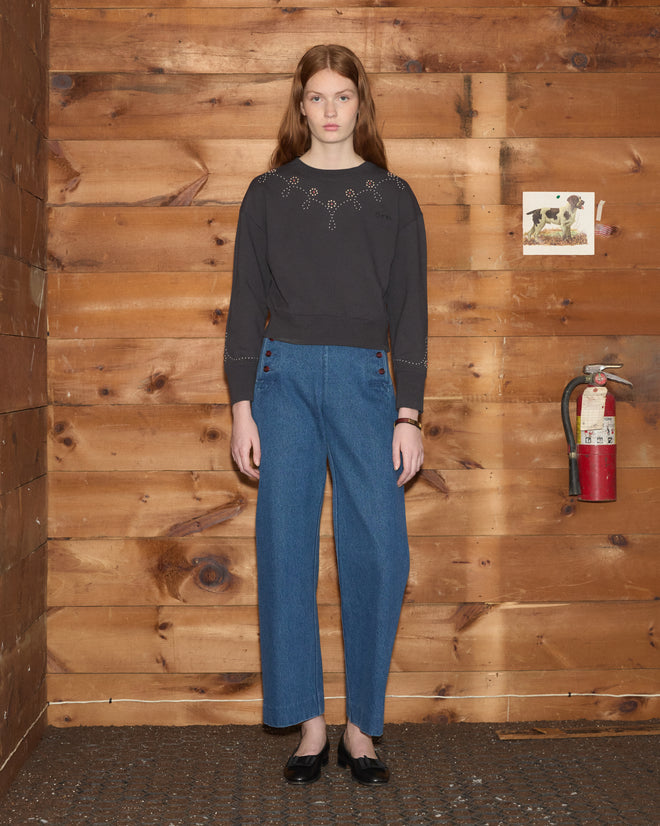 A woman with long hair wears a Silver Stud Sweatshirt and blue pants while standing by a wooden wall with a fire extinguisher.