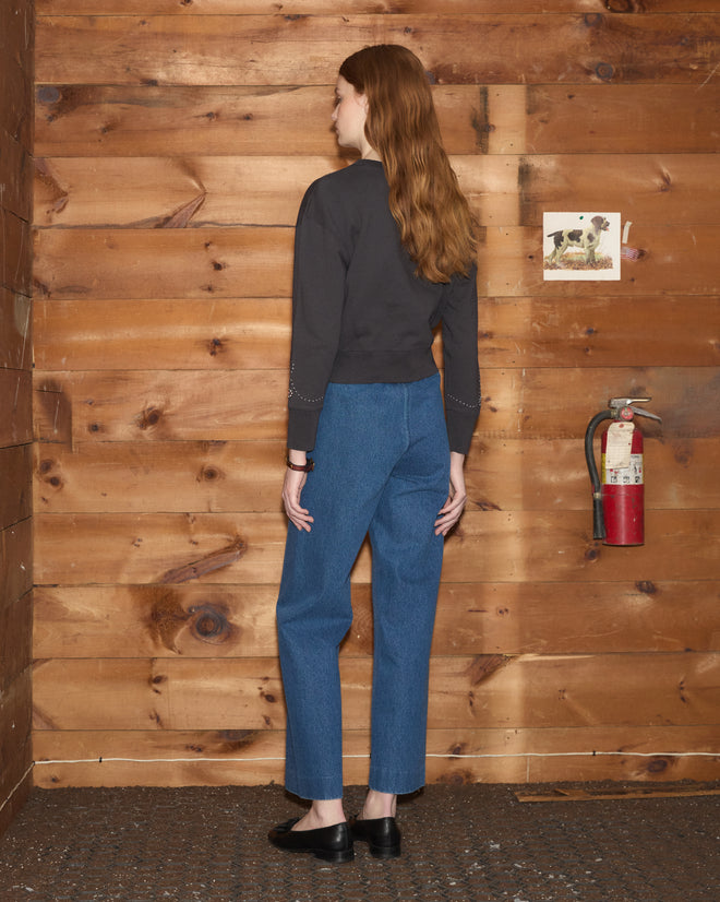 A woman with long hair stands facing a wooden wall near a fire extinguisher, wearing jeans, black shoes, and the Silver Stud Sweatshirt.