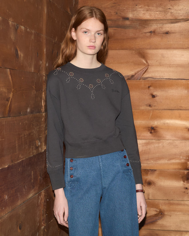 A person wearing the Silver Stud Sweatshirt and blue jeans stands by a wooden wall, looking at the camera.