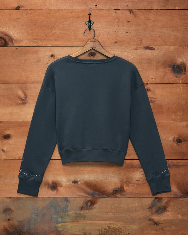 A Silver Stud Sweatshirt in dark gray soft cotton fleece, featuring gemstone-embellished sleeve cuffs, hangs on a wooden wall.