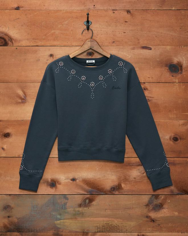 Silver Stud Sweatshirt—dark gray cotton fleece with small white floral patterns on the chest and sleeves, displayed against a wooden wall.