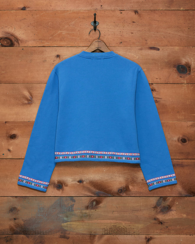 A Ribbon Cardigan in blue, featuring long sleeves, decorative trim, and antique pearl buttons, displayed against a wooden plank wall.