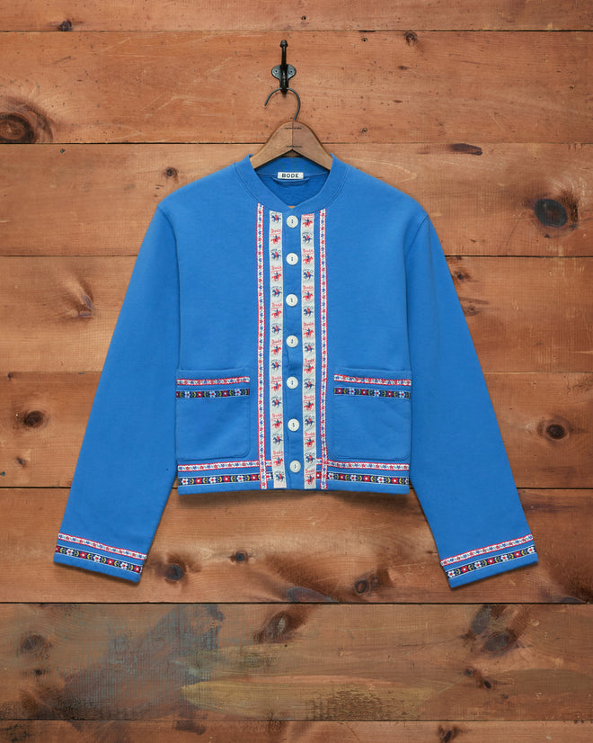 Ribbon Cardigan in blue, featuring decorative trim and antique pearl buttons inspired by 1960s Mod style, hangs on a hook against a wooden wall. Crafted from 100% cotton for enhanced comfort and timeless appeal.