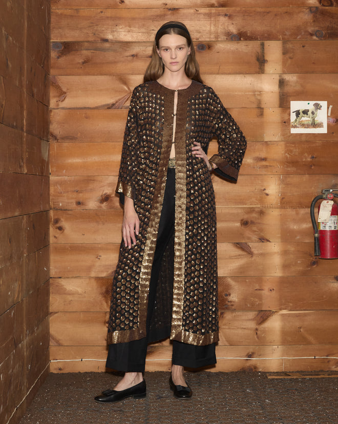 A woman wearing the Pyrite Sequin Kaftan stands in a wooden room near a fire extinguisher, exuding effortless elegance.