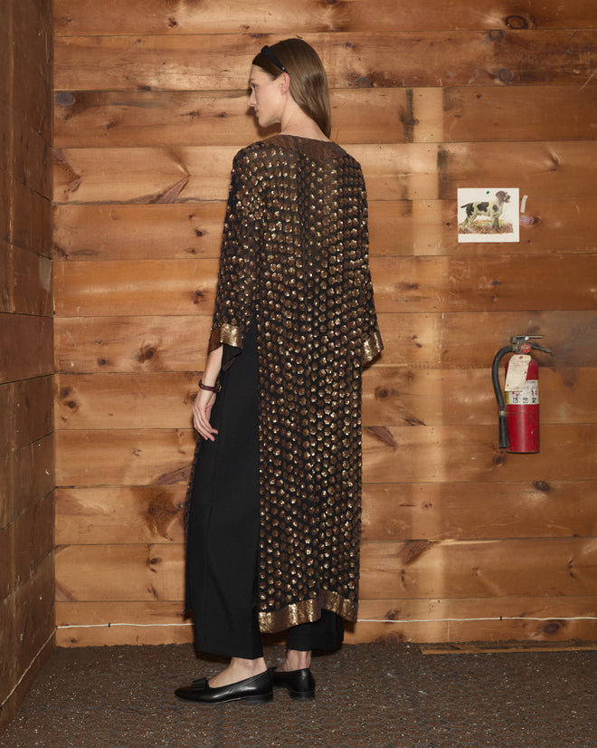 A woman in the Pyrite Sequin Kaftan—a long, dark coat adorned with gold patterns—stands with her back to the camera in a warmly lit wooden room.