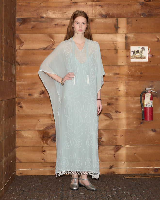 A woman wearing the Beaded Cirrus Kaftan—a pale blue silk garment adorned with delicate hand-beaded fringe—stands by a wood wall featuring a fire extinguisher and a small picture.