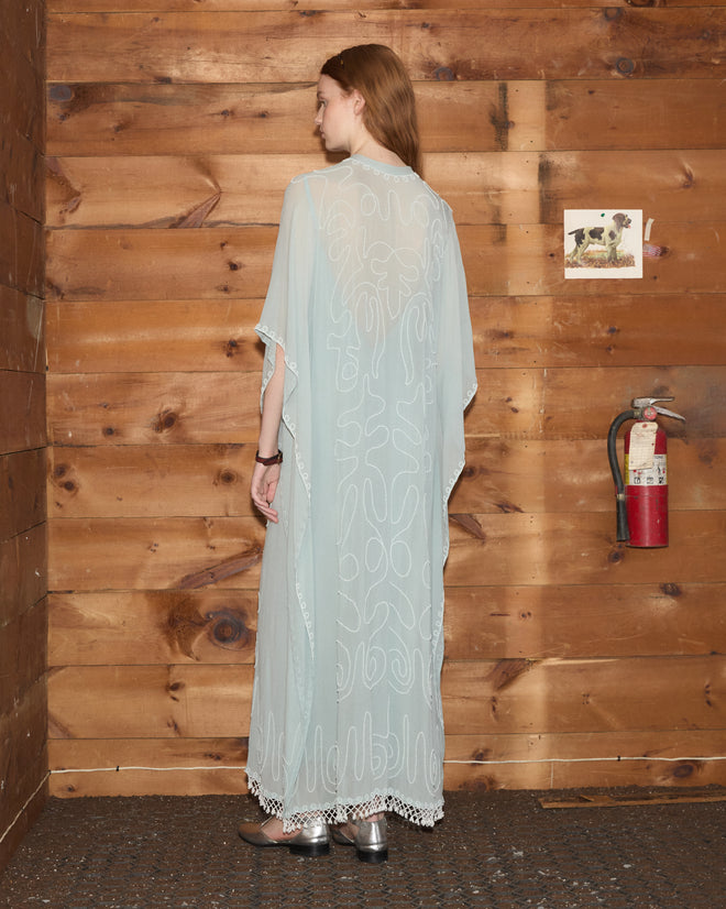 A woman in the Beaded Cirrus Kaftan, a light and sheer hand-beaded dress, stands with her back to the camera against a wooden wall with a fire extinguisher.