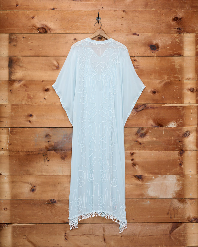 The Beaded Cirrus Kaftan, featuring light blue embroidered silk, loose sleeves, and a beaded fringe, hangs on a hook against a wooden wall background.