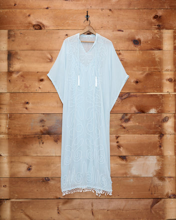 The Beaded Cirrus Kaftan, featuring sheer white silk with embroidered patterns and a delicate beaded fringe, hangs on a wooden wall.
