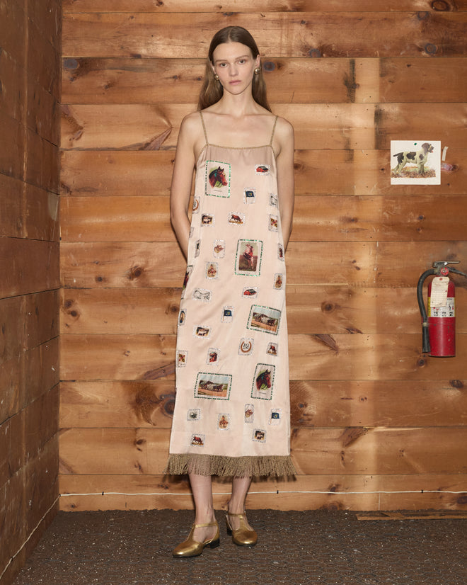 A woman stands indoors by a wooden wall and a red fire extinguisher, wearing the Equestrian Patch Dress—a beige reproduction dress crafted from collectible fabric swatches with patchwork designs reminiscent of tobacco silks.
