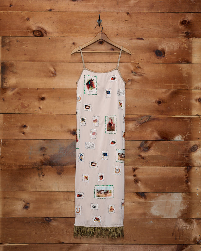 The Equestrian Patch Dress, featuring a beige slip silhouette with thin straps, patchwork imagery reminiscent of tobacco silks, and a fringe hem, hangs on a wooden wall.