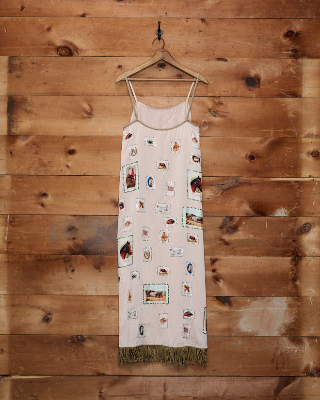The Equestrian Patch Dress, a sleeveless reproduction style featuring patchwork designs made from collectible fabric swatches and finished with a fringe hem, hangs on a wooden wall.