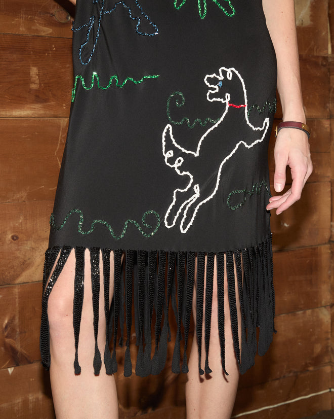 Dream Pony Dress—crafted from luxurious 100% silk crêpe de chine—features black beadwork with a beaded dog motif and a fringe hem; a person’s hand is visible against a wooden background.