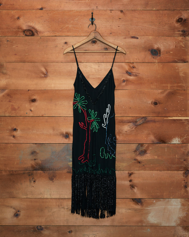 The Dream Pony Dress, crafted from luxurious 100% silk crêpe de chine and adorned with colorful palm tree designs and black fringe, hangs on a wooden wall.