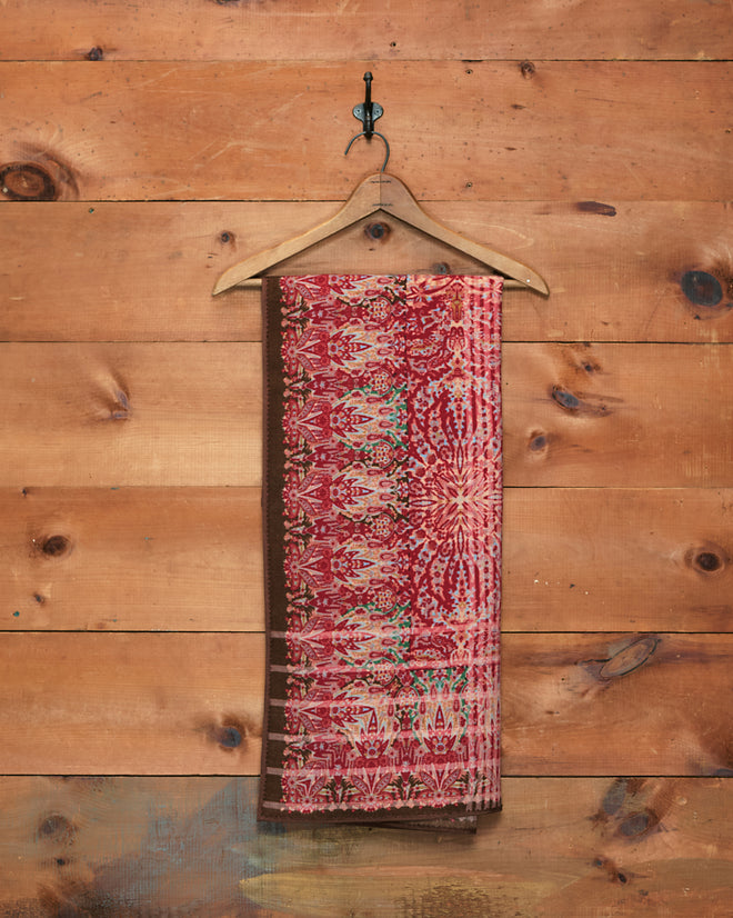 The Arrowleaf Scarf, crafted from soft wool in a red paisley pattern reminiscent of traditional Kashmiri shawls, hangs on a wooden hanger against a rustic wooden wall.