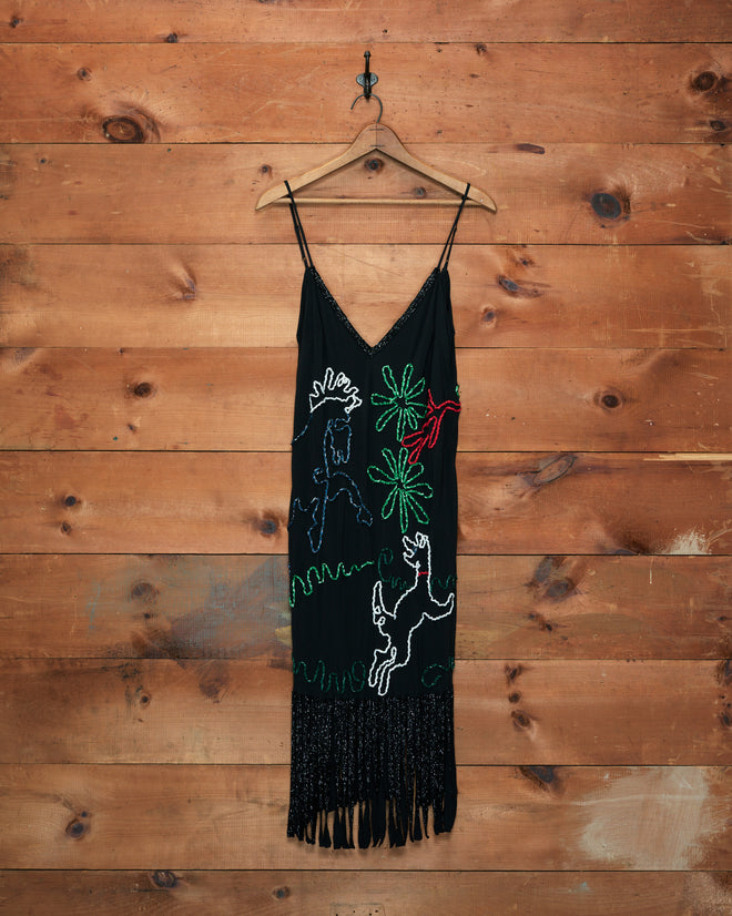 The Dream Pony Dress, crafted from black 100% silk crêpe de chine and featuring colorful animal and plant designs with fringe at the hem, hangs on a wooden wall.