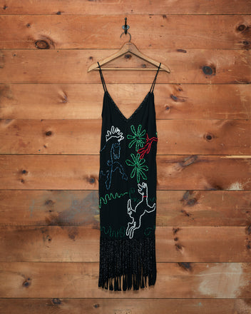 The Dream Pony Dress, crafted from black 100% silk crêpe de chine and featuring colorful animal and plant designs with fringe at the hem, hangs on a wooden wall.