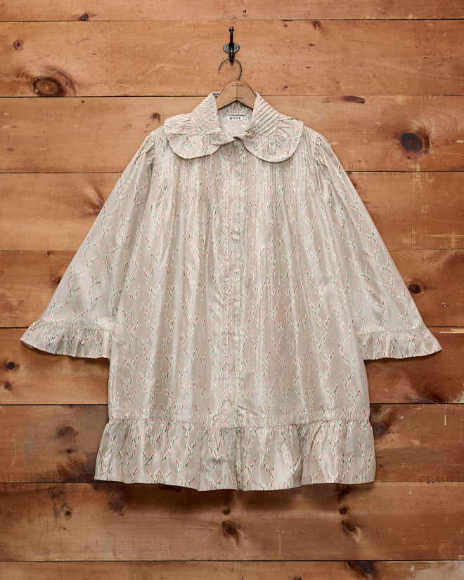 The Sagebrush Dress is a cream-colored, long-sleeved antique nightgown dress featuring a ruffled hem and an oversized Peter Pan collar, displayed hanging on a wooden wall.