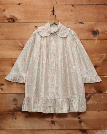 The Sagebrush Dress is a cream-colored, long-sleeved antique nightgown dress featuring a ruffled hem and an oversized Peter Pan collar, displayed hanging on a wooden wall.
