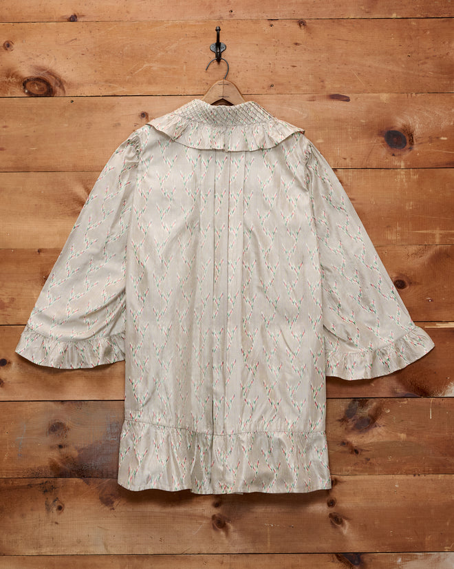 The Sagebrush Dress, a long-sleeved nightgown featuring a delicate light pattern and elegant ruffles, exudes timeless charm as it hangs on a wooden wall.