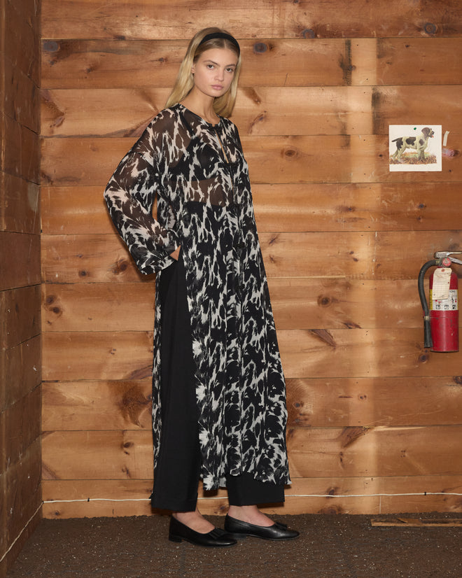 A woman wearing the Appaloosa Kaftan—a long, black-and-white silk piece inspired by the Appaloosa pattern—stands beside a wooden wall adorned with a fire extinguisher and a dog picture.