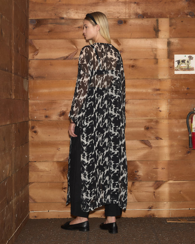 A woman wearing the long, black-and-white Appaloosa Kaftan stands sideways against a wooden wall indoors.