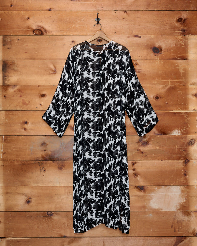 Appaloosa Kaftan – a black and white abstract patterned long dress with three-quarter sleeves, reminiscent of an Appaloosa pattern, hanging on a wooden wall.