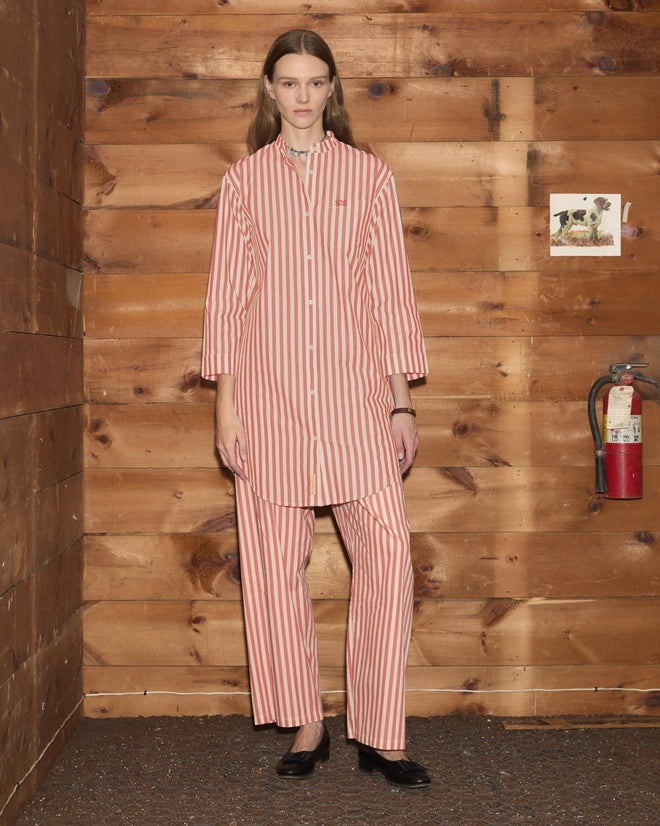 A person stands indoors by a wooden wall with a fire extinguisher and small photo, wearing the Rancho Tunic—a shirting-inspired, red and white striped top made from 100% cotton.