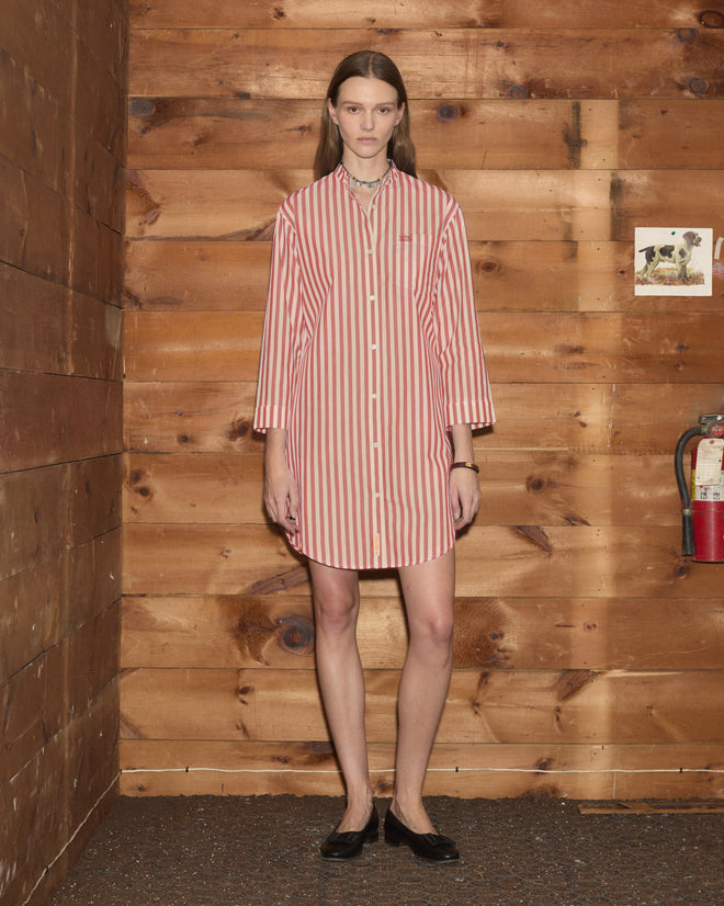 A woman stands against a wooden wall near a fire extinguisher, wearing the Rancho Tunic—a shirting-inspired dress with red and white stripes, crafted from 100% cotton.