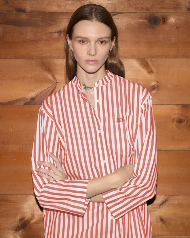 A person wearing the Rancho Tunic—a shirting-inspired, red and white striped shirt—stands with arms crossed in front of a wooden wall.