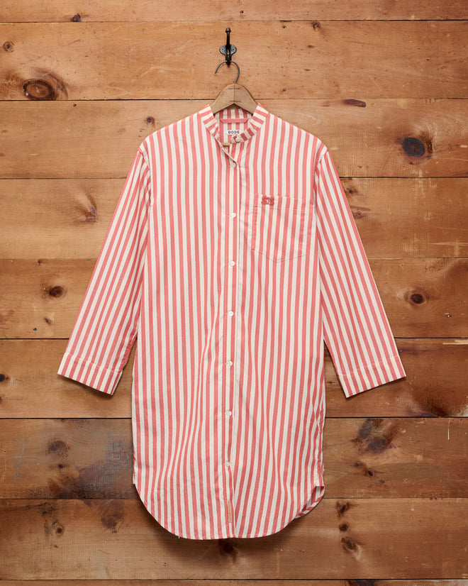 The Rancho Tunic, a long shirt with red and white vertical stripes and buttons, shirting-inspired and crafted from 100% cotton, is displayed hanging on a wooden wall.