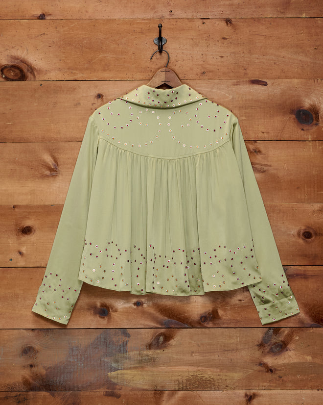 The Gem Dust Blouse, a light green long-sleeve Western-style blouse adorned with decorative studs, hangs on a hook against a wooden wall.