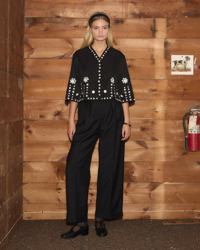 Woman wearing a black Pearly Royal Overshirt with pearl button embellishments and matching wide-leg trousers.