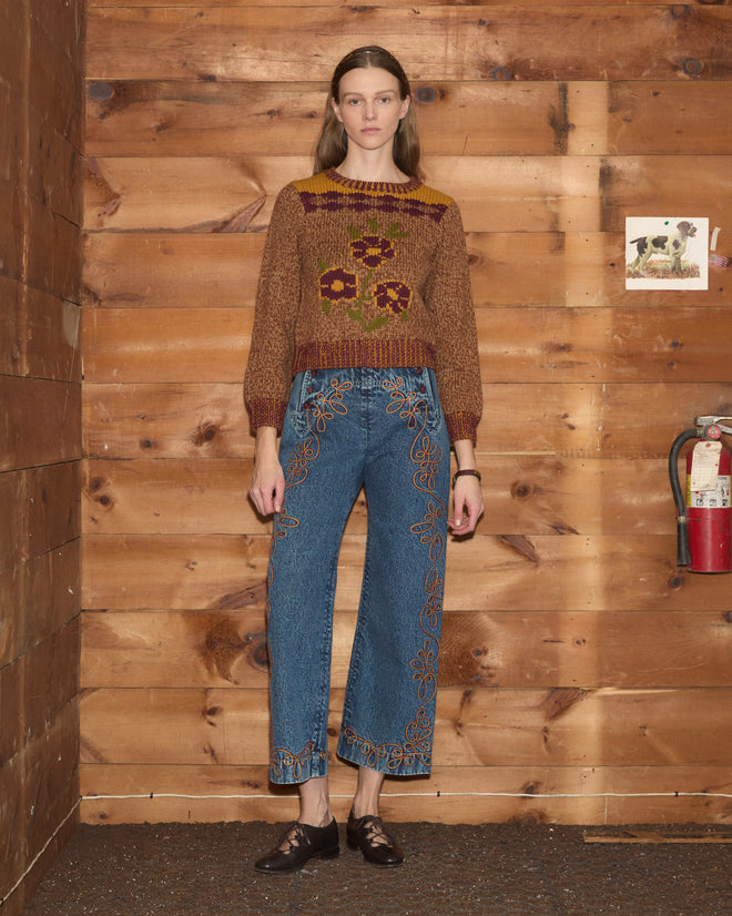 A woman in the Desert Rose Sweater, a shrunken brown floral knit crafted from soft Andean Highland wool, and blue embroidered jeans stands against a wooden wall with a fire extinguisher.