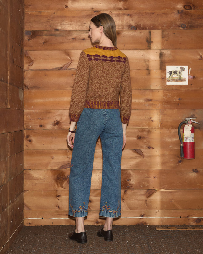 Woman wearing the Desert Rose Sweater and jeans stands facing a wood wall with a fire extinguisher and small photo.
