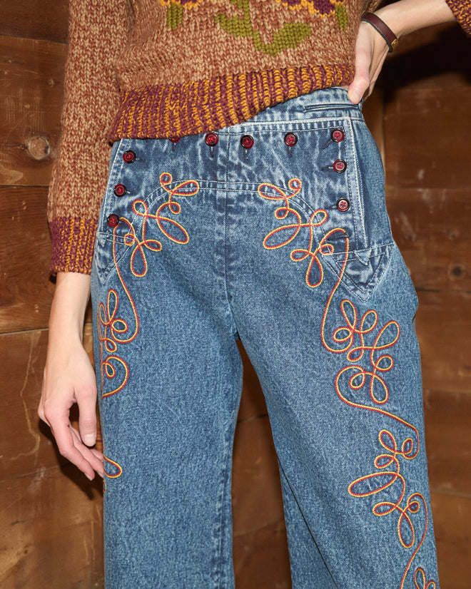 Rawlins Sailor Trousers featuring orange floral embroidery and red buttons, styled with a brown knit sweater and subtle cowboy-inspired accents, photographed against a wood wall.