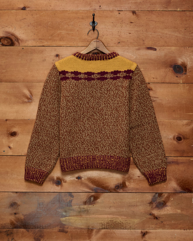 The Desert Rose Sweater, crafted from cozy Andean Highland wool in a brown knit and adorned with yellow and maroon patterns, hangs on a hook against a wooden wall.