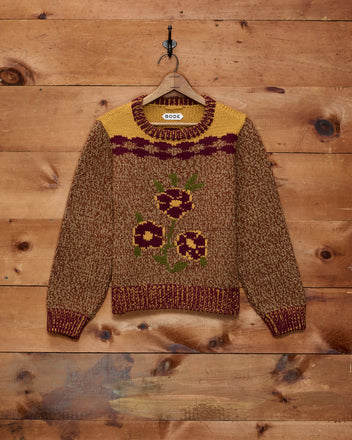 The Desert Rose Sweater, crafted from Andean Highland wool in a brown knit and adorned with yellow and maroon floral patterns, hangs on a wooden wall.