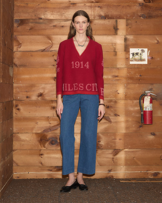 A woman wearing the Beaded Round-Up Sweater and blue jeans stands indoors by a wooden wall with a fire extinguisher and painting, her outfit evoking the vibrant spirit of the Miles City Round-Up.