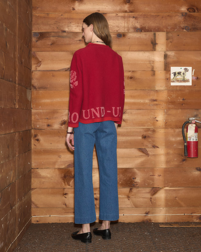 Wearing the Beaded Round-Up Sweater with blue jeans, a person stands indoors next to a wooden wall featuring a fire extinguisher and a photo of a dog.