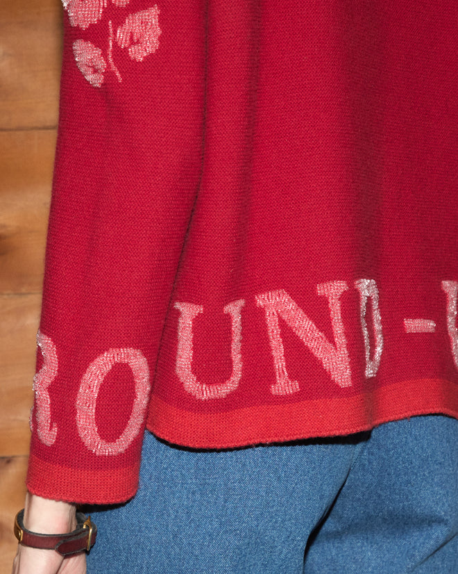 Wearing the Beaded Round-Up Sweater—a red top featuring “ROUND-UP” in white text—paired with blue jeans by a wooden wall, this look offers a stylish nod to the spirit of the Miles City Round-Up.