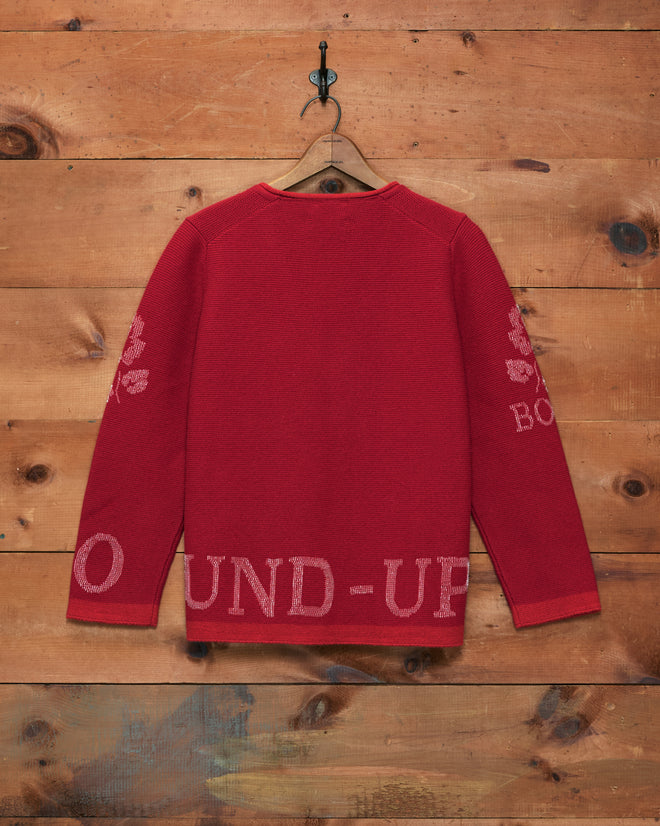 Beaded Round-Up Sweater in vibrant red, featuring light pink text and sleeve designs inspired by the classic Miles City Round-Up, displayed on a wooden wall with a hook.