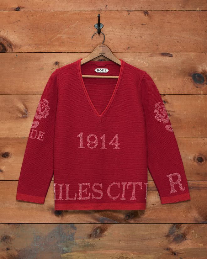 Beaded Round-Up Sweater in red with a V-neck and “1914,” “R,” and “RODE” text, inspired by the Miles City Round-Up spirit, displayed against a wood wall background.