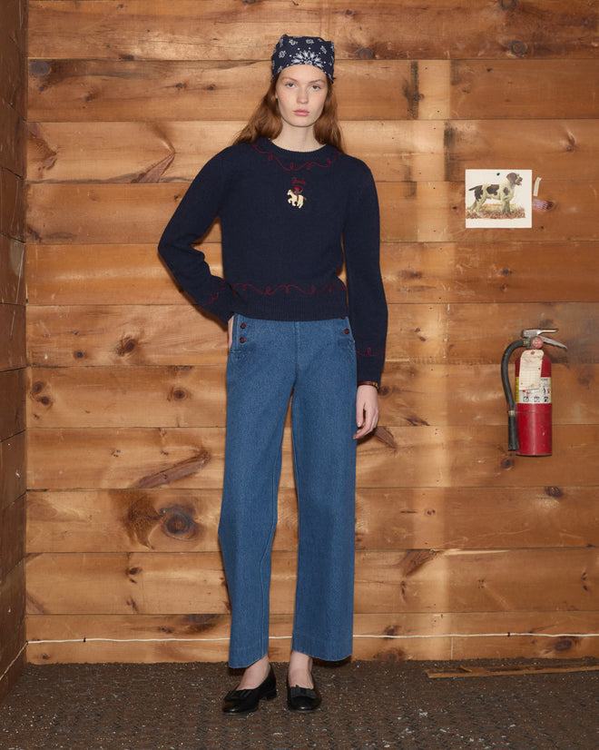 Woman in the Cowboy Appliqué Pullover and jeans stands indoors by a wooden wall with a fire extinguisher and photo.
