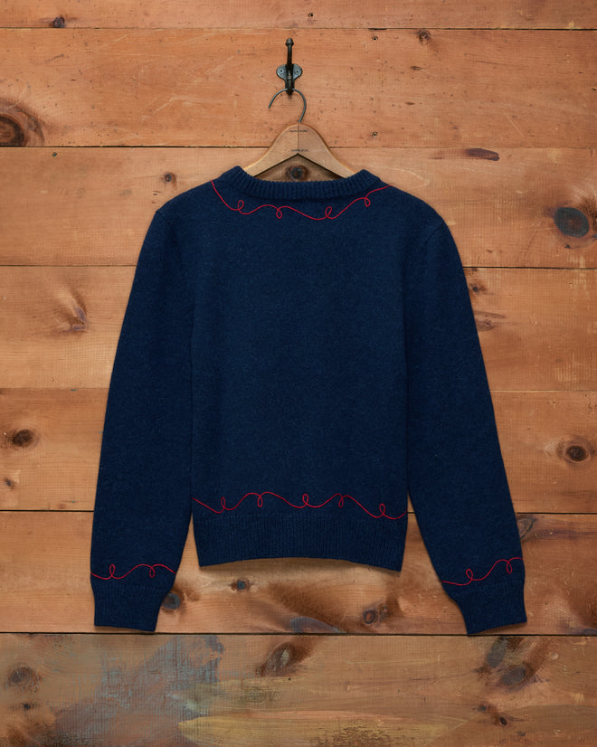 A Cowboy Appliqué Pullover in navy blue with red squiggly embroidery hangs on a wooden wall.