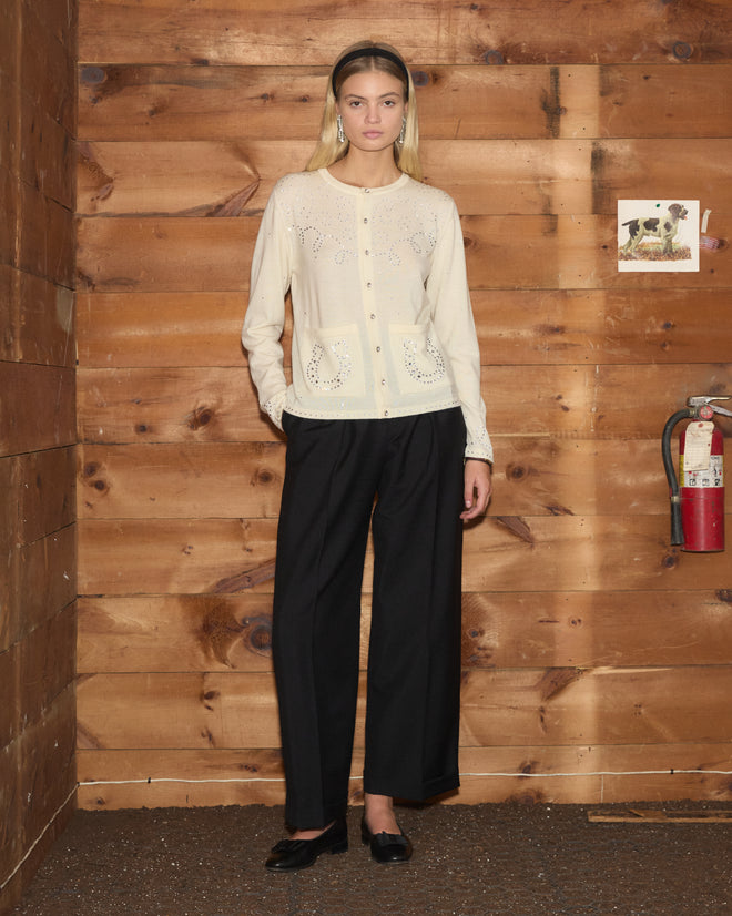 A woman wearing the Rodeo Rhinestone Cardigan and black pants stands by a wood wall with a fire extinguisher and a small dog photo.