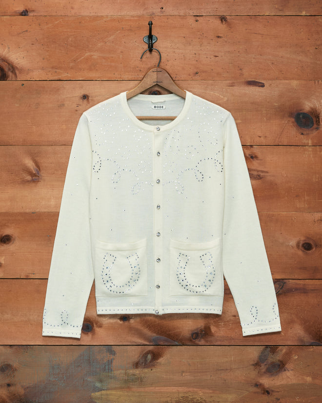 The Rodeo Rhinestone Cardigan is a cream merino wool sweater adorned with sparkling rhinestone horseshoe and swirl designs, displayed on a wooden wall background—making it the perfect horse motif piece to add a touch of glamour to your wardrobe.