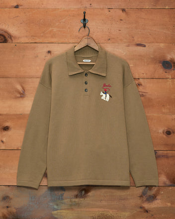 Cowboy Appliqué Polo - Tan, a long-sleeve merino wool shirt featuring a bear patch, displayed hanging on a wooden wall.