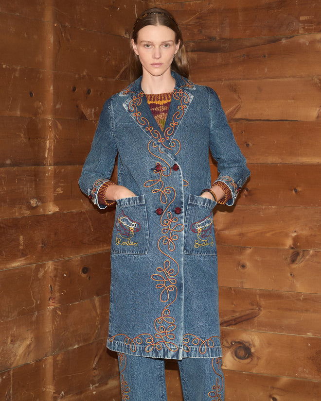 Wearing the Rawlins Coat, a woman stands against a wood-paneled wall with her hands in her pockets, channeling modern cowboy style with a touch of classic charm.